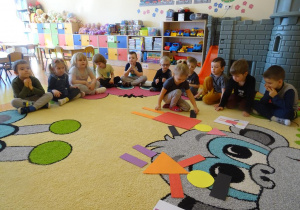 Dziewczynka układa pajaca z figur geometrycznych według wzoru, dzieci siedzą wokół niego i obserwują z zainteresowaniem.